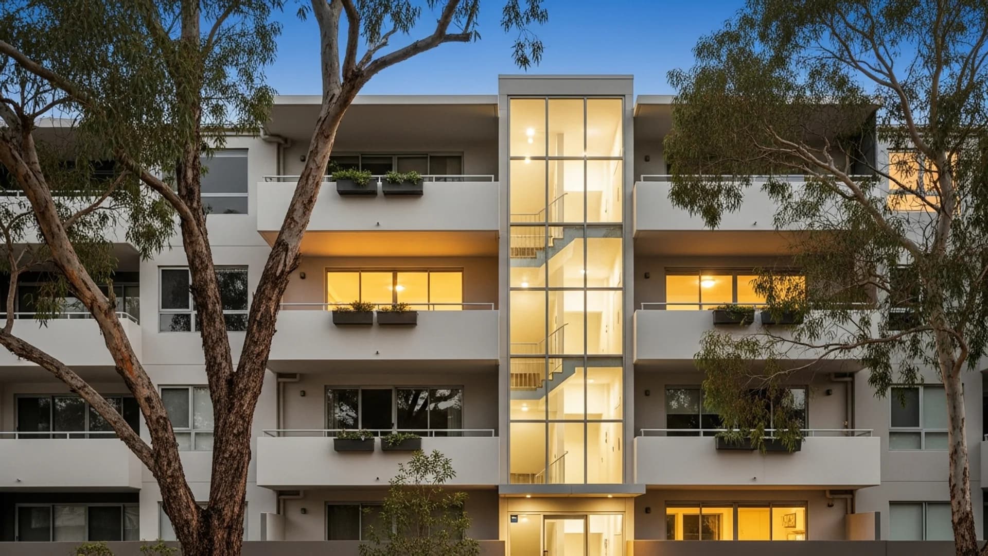 Modern Australian apartment building at dusk with a glazed stair-and-lift core lit warmly from within